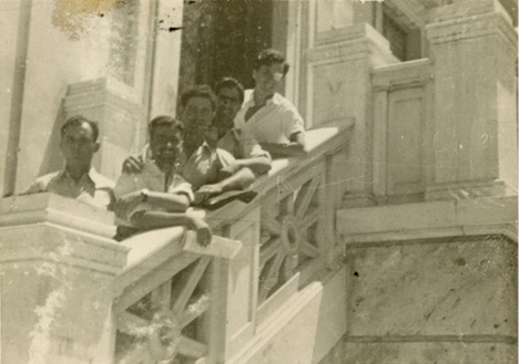 A vintage sepia-toned photograph shows five people standing on a marble staircase. They are leaning on the ornate railing, dressed in casual attire. The background features an elaborately designed building with decorative architectural elements.