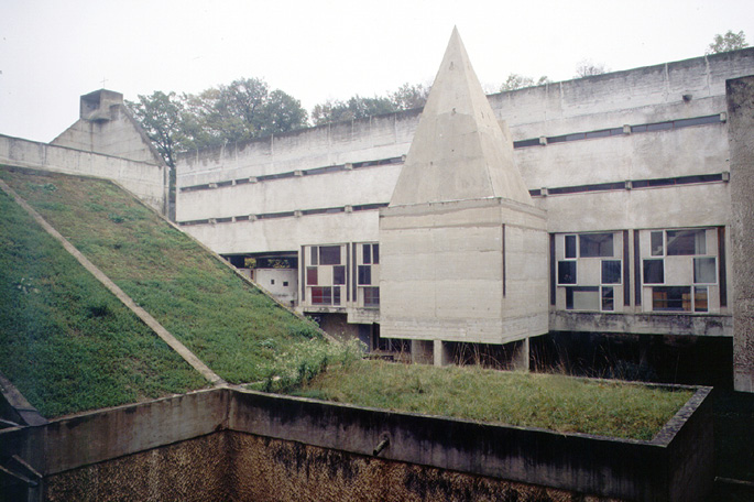 The image depicts a Brutalist architecture building with geometric shapes, featuring concrete structures and windows of varying sizes. There is a grassy rooftop area in the foreground, and a building with a prominent triangular structure in the center. Trees are visible in the background.