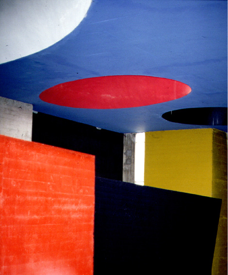 A corner of a concrete building consisting of large, colorful geometric shapes and a window cut-out. The image features red, yellow, black, and bluish-gray rectangular forms intersected with a blue ceiling and circular cutouts in red, white, and black.