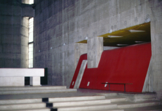 Interior of a modern concrete building with large red and white geometric structures. The space features exposed concrete walls, a set of stairs leading to an altar, and a mix of sharp and smooth angles. Light filters in through narrow vertical windows on the left.