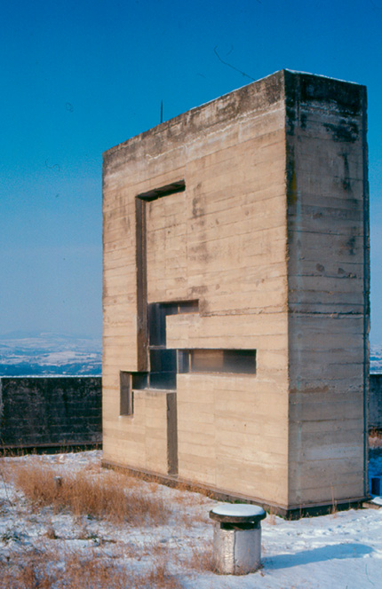 An abstract, rectangular concrete structure stands outdoors amidst a snowy landscape. The structure features various geometric cutouts and shapes embedded into its surface. The background shows a clear sky and distant, snow-covered hills.