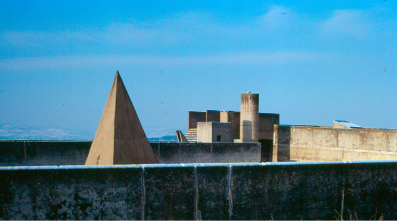 A concrete architectural structure featuring geometric forms, including a prominent pyramid and cylindrical tower, set against a clear blue sky. The surroundings include other concrete elements with a mountainous landscape visible in the distant background.