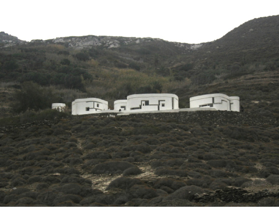A group of modern, white, cylindrical buildings sits perched on a rugged hillside with sparse vegetation. The buildings, featuring windows in abstract linear patterns, contrast against the earthy tones of the surrounding landscape and the mountainous backdrop.