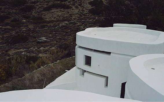 A modern, white, cylindrical building with a flat roof, featuring minimalist, rectangular windows and rounded edges. The structure is set against a natural backdrop with rocky terrain and sparse vegetation under cloudy skies.