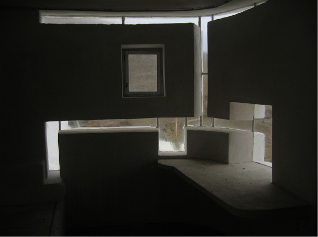 A dimly lit room with an abstract architectural design featuring irregularly shaped white walls and several small windows letting in natural light. A ledge extends from the wall, forming a unique corner seating or display area. The scene is minimalist.