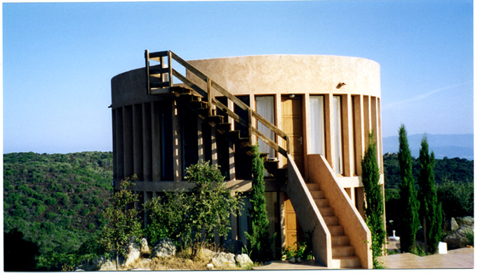 A light brown, cylindrical building with exterior stairs to the first floor and the roof, with long rectungular windows running round the perimeter at regular intervals. The building is perched on a hillside with small green trees.