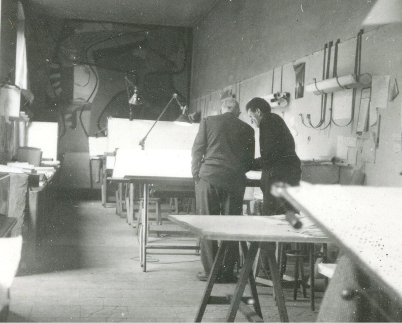 Black and white photo of two people standing and discussing something on a large drafting table in a room filled with drafting equipment. The room has long desks, lamps, and various papers pinned to the walls. The atmosphere is academic and studious.