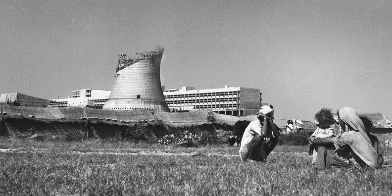 Black and white photo of three people in Indian rural dress seated on grass with woven mats next to a child and a cow. A distinctive cone-shaped structure under construction is in view, in front of several buildings. The sky is clear with no clouds.