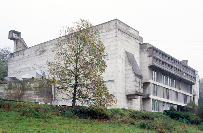 A large, Brutalist-style concrete building with minimal windows and a distinct angular design stands on a grassy hillside. A single tree with sparse leaves is in the foreground. The sky is overcast, and there are vents and structures visible on the building’s roof.