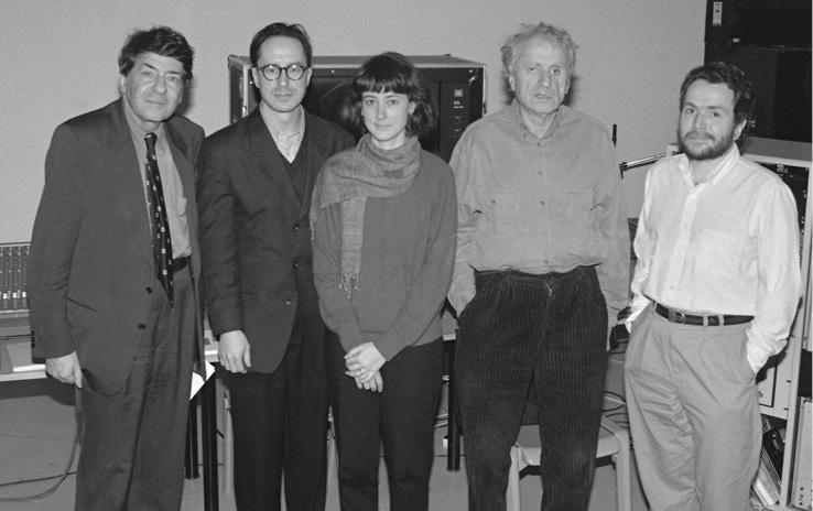 A black-and-white photo features a group of four men standing side by side with one woman in the middle. They are dressed in business-casual attire, with a mix of formal wear and sweaters. Behind them is various items of recording equipment.