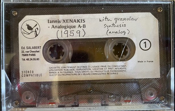 A close-up of an analog cassette tape in a clear plastic case. The label reads “Iannis Xenakis - Analogique A-B (1959)” with additional handwritten text “with granular synthesis (analog)” on the label. Various other small printed details are visible.