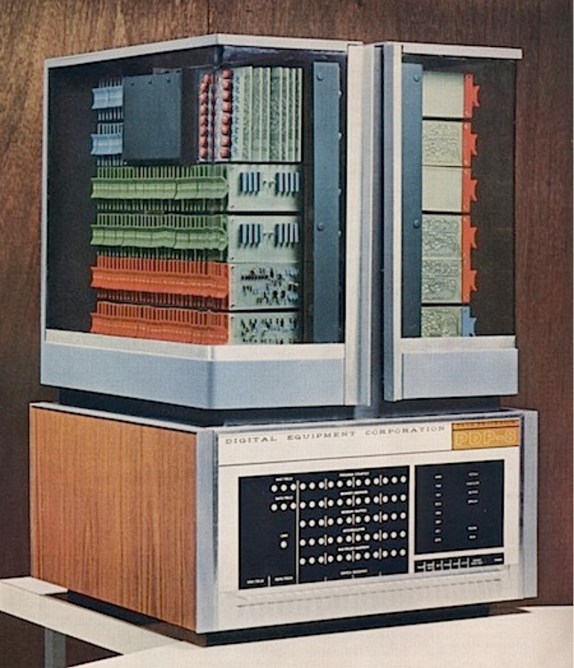 A vintage PDP-8 minicomputer from Digital Equipment Corporation is shown. The computer features an open top displaying colorful circuit boards and a control panel at the bottom with numerous switches and lights. The case is made of brushed metal and wood panels.