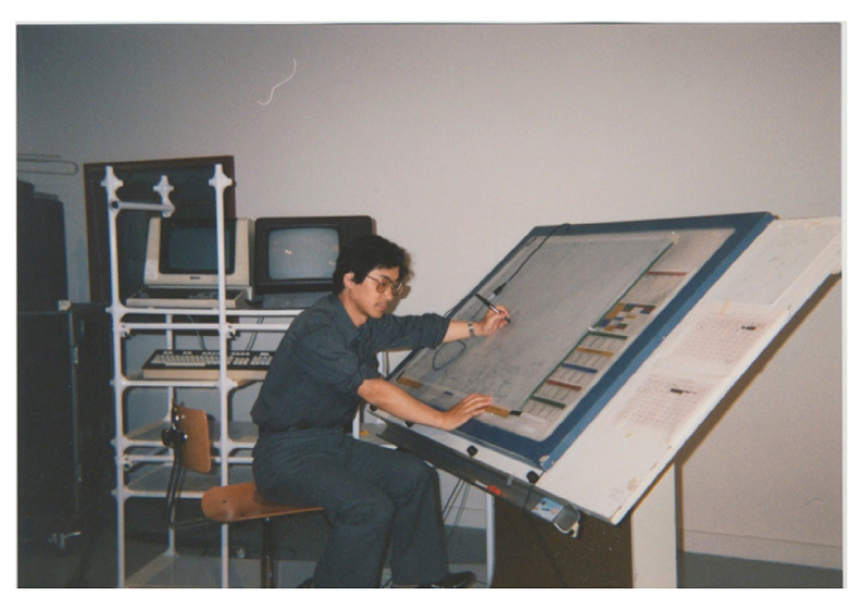 A person sits at the large drafting table of the UPIC, working with a stylus on the drawing surface. Behind them, there are vintage computer monitors and equipment on shelves. The person appears focused on their task in a workspace setting.