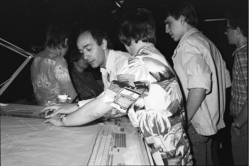 A black-and-white photo shows a group of five men gathered around the large drafting table of the UPIC, engrossed in a collaborative effort. The man in the foreground points at the table, which is covered with papers and drafting tools. The scene is set in a dimly lit room.