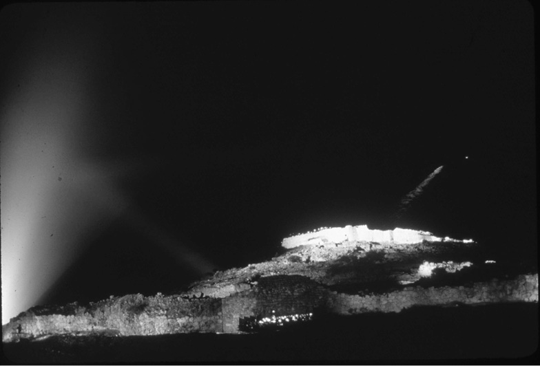 A night view of ancient ruins illuminated by lights. The crumbling stone structures are partially bathed in warm light, while beams of blue light can be seen piercing the night sky from the left side of the image. The scene has a dramatic and mysterious atmosphere.
