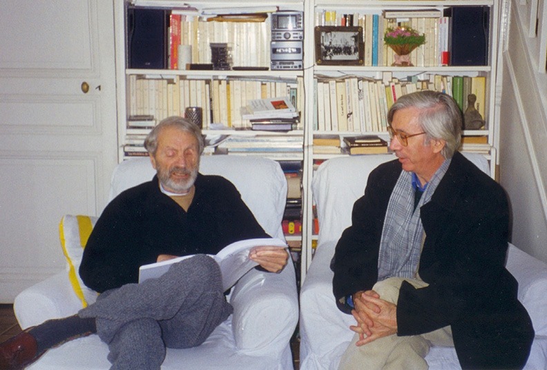 Two older men are seated on white armchairs in a cozy room with a bookshelf filled with books and a few decorative items in the background. The man on the left is Xenakis reading a book out loud, while the man on the right, Candido Lima is listening attentively, with his hands clasped.