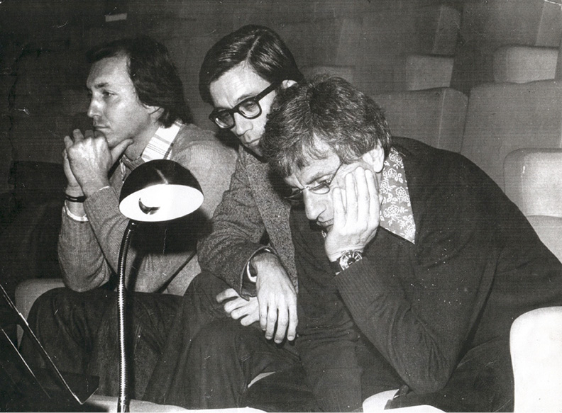 Three men are sitting closely together, looking thoughtful and focused. Two of them are leaning forward with their hands near their faces. There is an illuminated desk lamp in front of them, casting light onto papers they are observing. The background is dimly lit.