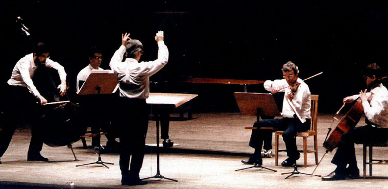 A music ensemble including three performers and a pianist performs on stage. The bassist stands to the left, bent over, while the other three musicians play seated. The conductor, standing at a podium, raises his hands. All are dressed in white shirts and dark pants, with music stands in front of them.