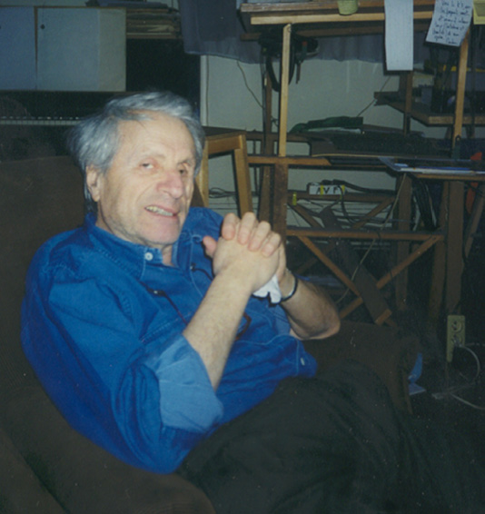 Iannis Xenakis with grey hair, dressed in a blue shirt and black pants, sits comfortably on a brown couch. He is smiling and clasping his hands. In the background, there is a cluttered workspace with papers, books, and a desk.