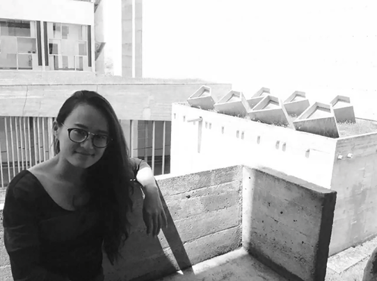 Yin Yu with long hair and glasses is sitting on a concrete ledge in an sunlit urban setting. She leans casually against a low wall, with modern buildings in the background. The image is in black and white.