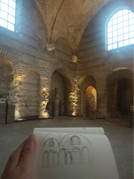 A person holds a sketchbook with a drawing of an ancient architectural structure. The drawing mirrors the interior of the historic stone building they are inside, featuring arched windows, doorways, and illuminated details on the walls and ceilings.