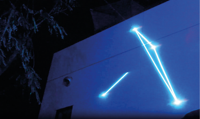 A building’s wall illuminated with bright blue laser beams forming geometric patterns at night. Part of a tree is visible on the left side of the image. The scene has an ethereal and futuristic appearance.