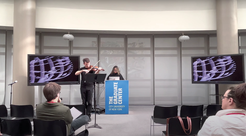 A violinist and a pianist perform on a stage at The Graduate Center, City University of New York. The violinist is standing and playing, while the pianist is seated at a podium keyboard. Large screens displaying abstract patterns flank both sides of the stage.