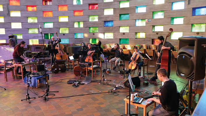 A group of musicians performs in a brightly lit room with multicolored windows. The ensemble includes cellos, double basses, and electronic music equipment. They are surrounded by an array of microphones, cables, and sound equipment, creating an eclectic atmosphere.