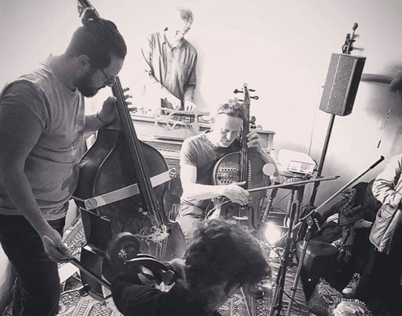Black and white photo of a small music performance in a cozy, dimly-lit room. Three musicians are playing string instruments passionately, while one person operates a mixing board. Various microphones and equipment are set up around them.