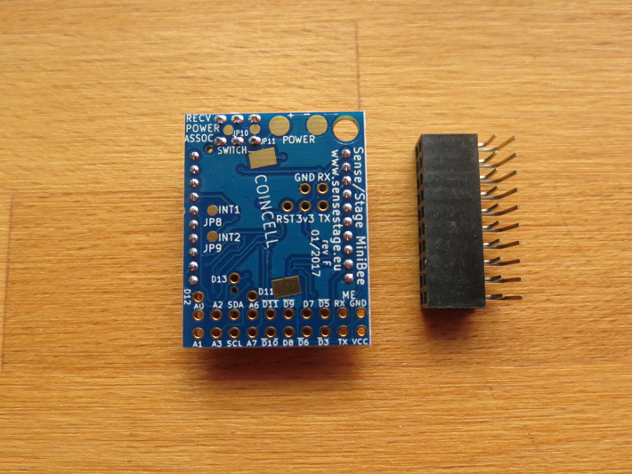 A small blue electronic circuit board with various labeled pins and components, including a cluster of holes and a switch. Next to it is a black, rectangular connector with multiple metal pins, both placed on a wooden surface.