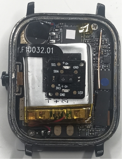 The image shows the internal components of a smartwatch with the back cover removed. Visible are the battery, circuit board, and various electronic parts, including a microcontroller and wiring, all compactly arranged within the watch casing.