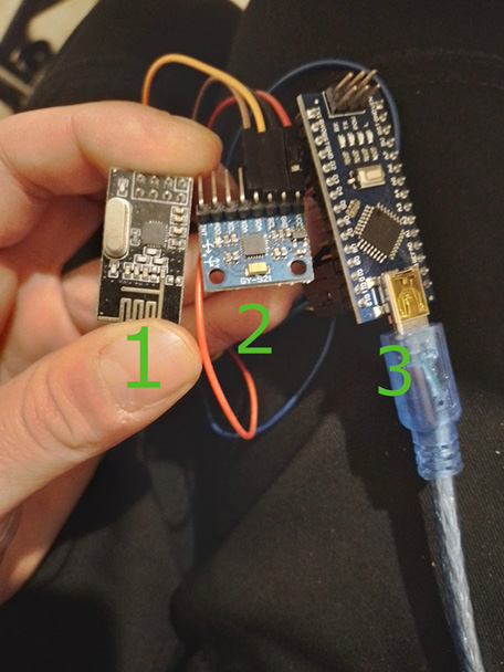 A hand holds three small electronic components labeled with green numbers. Component 1 is black and rectangular. Component 2 is blue with several pins and wires. Component 3 is a larger blue board with a USB cable attached.