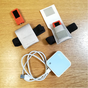A photo showing two small rectangular devices with black screens, each placed in a fabric pouch with Velcro straps, and a white USB cable coiled around a small, square, light blue device with the TP-Link logo. All items are on a wooden surface.