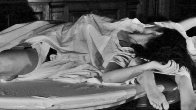 A person, partially covered by a sheet, lying on a mattress with their legs and one arm exposed. The setting appears dimly lit and informal. The person’s face is partially obscured by their arm and hair.