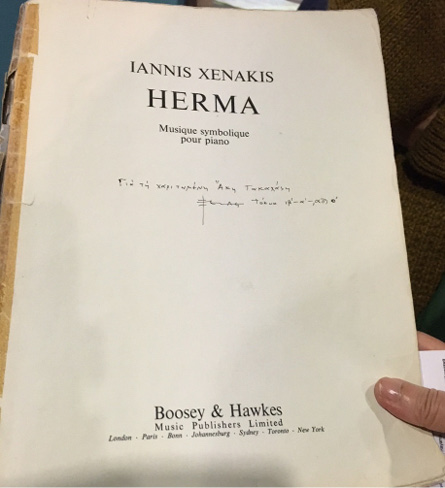 A person holds a music score titled “Herma” by Iannis Xenakis. The cover reads “Musique symbolique pour piano” and features handwritten notes in Greek, including a dedication to Aki Takahashi. Published by Boosey & Hawkes, the page is slightly worn at the edges, suggesting frequent use.