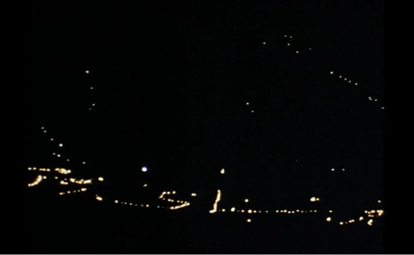 A night scene shows scattered lights from houses and street lamps, arranged in a curved, hilly pattern. The lighted areas form a meandering line across the dark background, suggesting a mountainside village or rural area under a night sky.