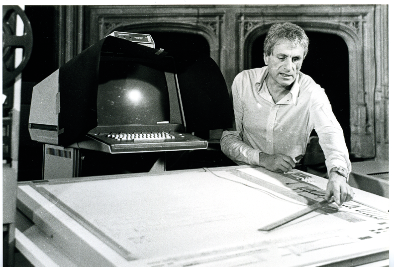 Iannis Xenakis in an open-neck shirt holding a plastic ruler leans over a large drawing board, which is part of the first version of his UPIC system. Next to him is an old-fashioned computer monitor with keyboard.