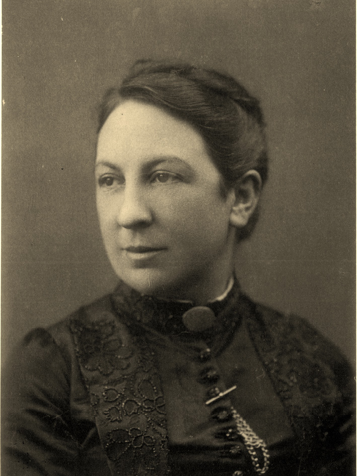 Photograph of a white woman, head-and-shoulders, dark hair pinned up. Ornate black dress with buttons and a watch chain.
