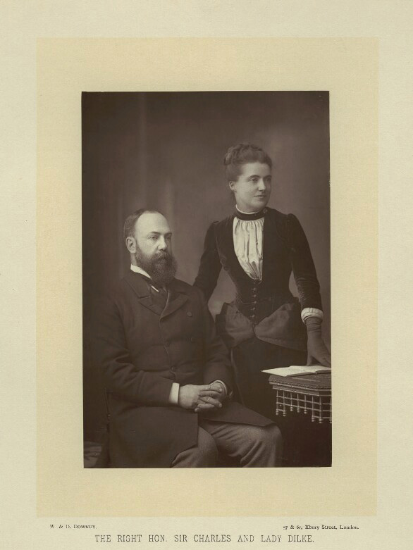 Photograph of a seated man and standing woman, both middle-aged. He has receding hairline, short beard, frock coat. She has dark hair, pinned up and black dress with white fichu.