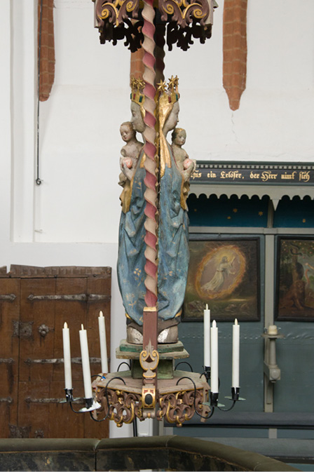 The candelabrum is seen from the level of the nuns’ choir above the dark-brown wooden screen dividing it from the east side of the church. The double figure of the crowned Mary, clad in a red tunic and a blue coat and holding the naked Christ Child, is surrounded by the golden rays of the sun; she stands on the silver face of the moon. Above her is a carved wooden baldachin painted in dark red with gold.
