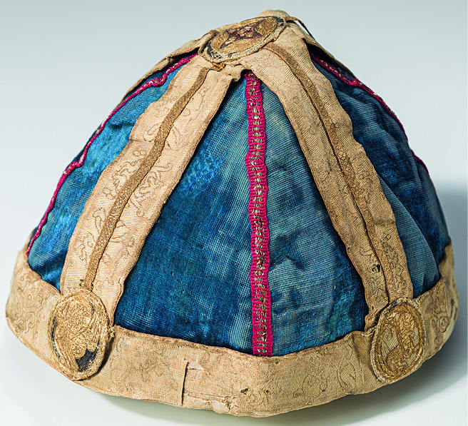 The nuns’ crown consists of two cross-bands attached to a circlet, all woven through with gold and embellished with embroidered motifs. Five textile medallions are applied at the points where the bands meet, of which three are visible in the photograph: a cherub on the left and the Lamb of God. The more complex embroidery technique and more luxurious threads used for the Lamb indicate that it would have been positioned at the front, worked in gold on a blue ground. At the apex is an indented diamond in red thread outlined in blue and punctuated with gold circles on a gold ground of couched embroidery. The crown is sewn onto a blue velvet cap of later date as a support.