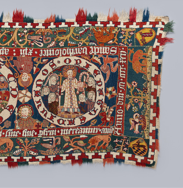 Detail from a tapestry with the life of Bartholomew, featuring the last scene on the right end of the textile. The background is embroidered in blue, the text in contrasting red and white thread. In the roundel, Bartholomew is portrayed as a young man with a crown and white garment; he carries his flayed skin, here depicted as a miniature figure hanging over his left arm. In his right hand he holds a cross. Surrounding him are six kneeling nuns, their hands raised in prayer. On the border strip a pelican plucks at its breast to feed its young and a lion roars over its cubs; they alternate with stylized trees. In the corners are coats of arms. For the interpretation and the text in the bands which surround this scene and run along the whole tapestry see the translation and discussion in Chapter III.2.