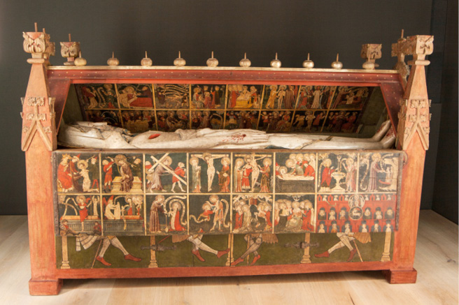 The photograph shows the Holy Sepulchre of Wienhausen with the front top lid opened to reveal the larger-than-life figure of Christ, painted white with the wounds highlighted in red. On the inside of the coffin, the wooden panels are decorated with scenes from the life of Christ. The front shows the biblical sequence from the flagellation to heavenly Jerusalem, with a focus on the resurrection and Christ’s appearances after it, such as Mary Magdalen’s encounter with Christ in the garden and Thomas’s putting of his hand into the wound in Christ’s side.