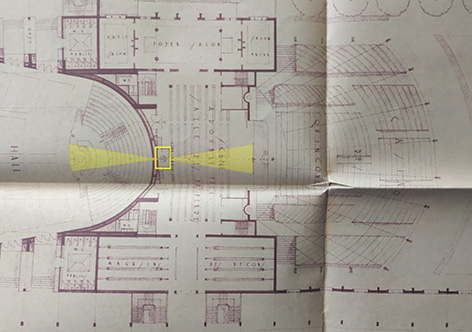 On voit la partie de l’intérieur du Palais Municipal dédiée aux spectacles, avec de gauche à droite : un grand hall d’exposition, un écran de cinéma qui lui tourne le dos, un gradin face à l’écran, une cabine de projection, une scène de théâtre (avec de petits dessous et des cintres réduits), et à nouveaux des gradins mais en extérieur.