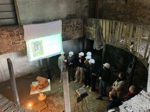 La photographie présente un point de vue en plongée sur le parterre du Vieux Théâtre abandonné, avec des étudiants présentant leurs recherches sur cet objet architectural devant les élus et propriétaires des lieux. En guise de support de présentation, un drap suspendu reçoit une vidéo-projection et une maquette en carton recompose la volumétrie originelle.
