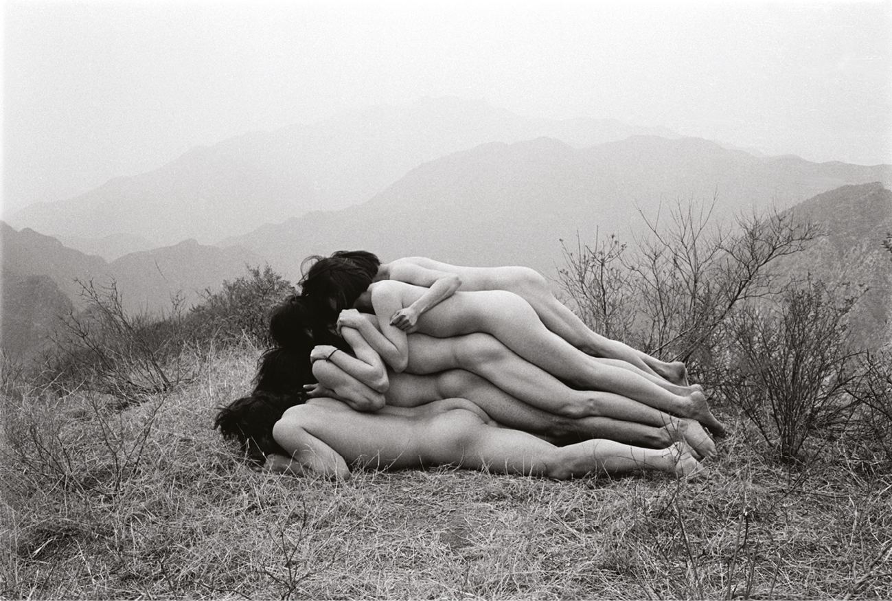 A black-and-white photograph of a performance art piece. In the image, several nude bodies are stacked on top of each other, lying horizontally in a pile on dry grass, as if forming a human mound. The performance takes place outdoors, with barren, rocky hills in the misty background, creating a serene yet stark contrast between the natural landscape and the intimate human arrangement. The bodies are positioned tightly together, contributing to the artist’s conceptual intent of ‘adding one meter’ to the mountain.