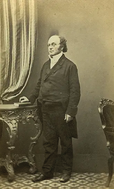 A sepia full-length photograph of a man in Victorian clothes standing next to an ornamental table and curtain.