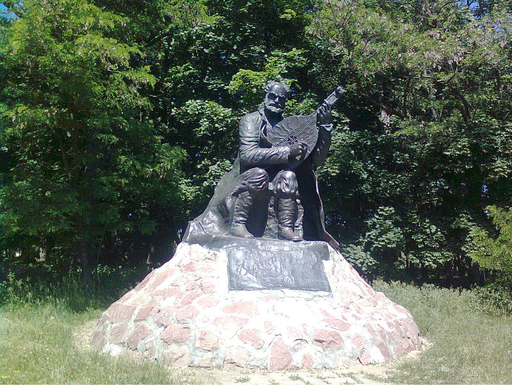 A photograph of the statue of an elderly man with a beard, playing a traditional Ukrainian bandura, wearing simple rustic clothing.