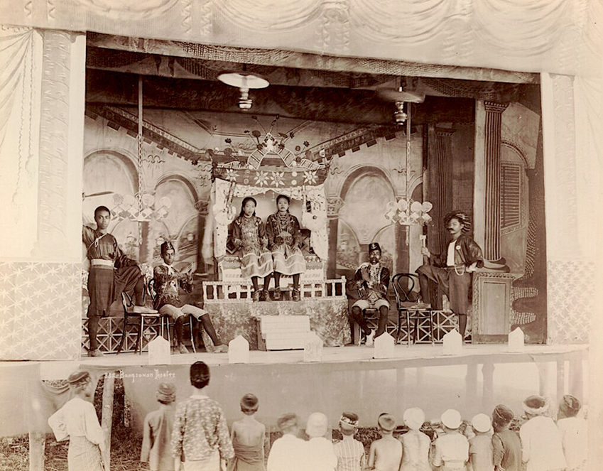 A historical photograph of a traditional Bangsawan theatre performance, showing actors in elaborate costumes on a decorated stage with an audience watching.