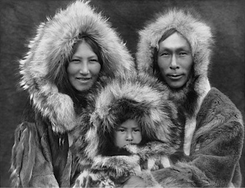 A historical black-and-white photograph of an Inuit family dressed in thick fur parkas. The mother and father smile gently while their young child, also wrapped in fur, gazes directly into the camera.